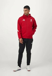 Sarkans sporta jaka ar Liverpool FC logo, baltas akcenti, kopā ar melniem sporta biksēm un baltām sporta kurpēm, stāvot uz vienkārša fona.