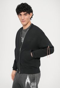 Sudadera negra con cremallera, bolsillos laterales, puños acanalados y un cordón decorativo en la manga izquierda. Llevada sobre una camiseta gris.