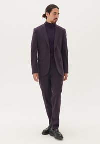 Lila öltöny, slim fit szabással, részletgazdag gallérral és két gombbal, amelyhez illő garbót és fekete cipőt viselnek. Sima textúra.