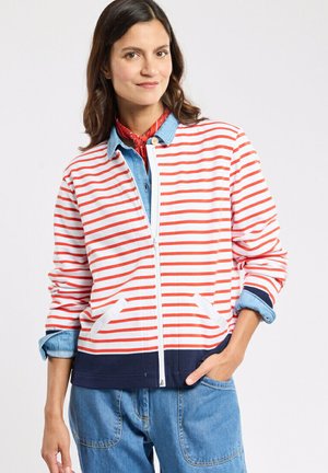 Femme portant une veste zippée rayée rouge et blanche sur une chemise en denim, avec un jean bleu et un foulard rouge au cou, debout avec une main dans la poche.