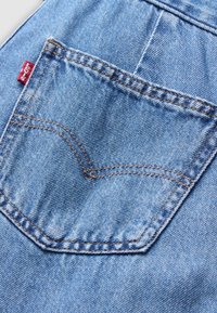 Μπλε υφαντό τζιν με πίσω τσέπη, έντονη πορτοκαλί ραφή και κόκκινη ετικέτα λογότυπου κοντά στο πάνω μέρος. Η υφή φαίνεται μαλακή και φθαρμένη.