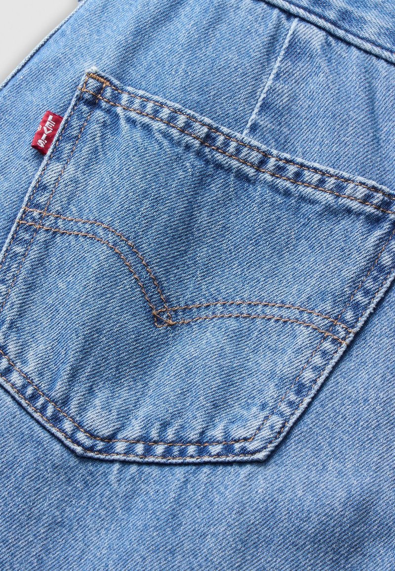 Tissu en denim bleu avec une poche arrière à surpiqûres orange contrastantes et une étiquette logo rouge près du haut. La texture semble douce et usée.