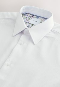 Chemise blanche avec col pointu, trois boutons visibles et doublure intérieure du col à motif floral avec étiquette de marque "NEXT" et étiquette de contenu en coton.
