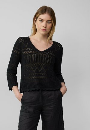 Schwarzer gestrickter Pullover mit V-Ausschnitt, drei Viertelärmeln, strukturierten Mustern und gewelltem Saum. Kombiniert mit lockeren schwarzen Hosen.