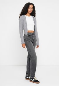 Γυναίκα με μακριά σγουρά μαλλιά, ντυμένη με γκρι ζακέτα με φερμουάρ, λευκό κοντό τοπ, ξεθωριασμένα μαύρα τζιν και μαύρα και άσπρα αθλητικά παπούτσια, που στέκεται σε λευκό φόντο.