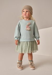 Hanorac verde mentă cu un patch cu iepure, purtat peste o fustă din tull floral deschis. Căciulă tricotată bej și adidași bej în două nuanțe cu accente de blană.