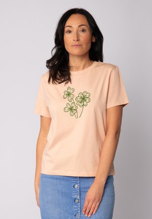 Pfirsichfarbendes Baumwoll-T-Shirt mit rundem Ausschnitt, kurzen Ärmeln und einer Illustration von drei grünen Kleeblättern auf der Vorderseite.