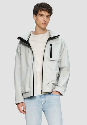 Veste gris clair avec capuche, accents noirs, fermeture éclair sur le devant et poche poitrine zippée. Fabriquée dans un matériau lisse et imperméable.