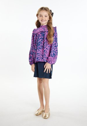 Lila und rosa Leopardenmuster Bluse mit langen Ärmeln und Rüschen, kombiniert mit einem marineblauen Rock und goldenen Slippers.