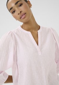 Chemise rose clair en tissu texturé avec un décolleté en V et des manches bouffantes. Motif à pois subtil sur l'ensemble. Design simple avec des accents minimalistes.