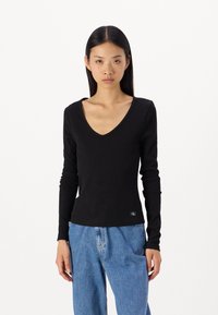 Μαύρο μακρυμάνικο τοπ σε σχήμα V φτιαγμένο από ριπ ύφασμα, με εφαρμοστή σιλουέτα και μια μικρή ετικέτα λογότυπου στο τελείωμα. Συνδυάζεται με φαρδιά μπλε τζιν.