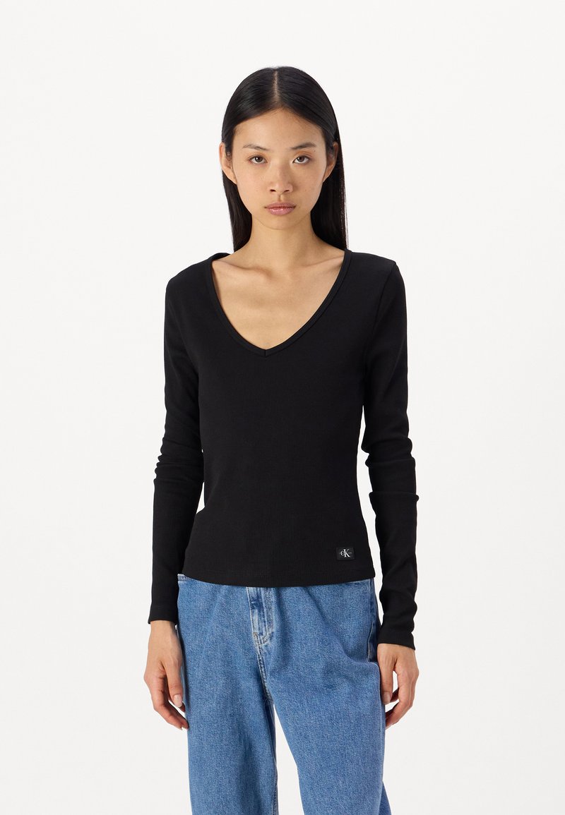 Μαύρο μακρυμάνικο τοπ σε σχήμα V φτιαγμένο από ριπ ύφασμα, με εφαρμοστή σιλουέτα και μια μικρή ετικέτα λογότυπου στο τελείωμα. Συνδυάζεται με φαρδιά μπλε τζιν.