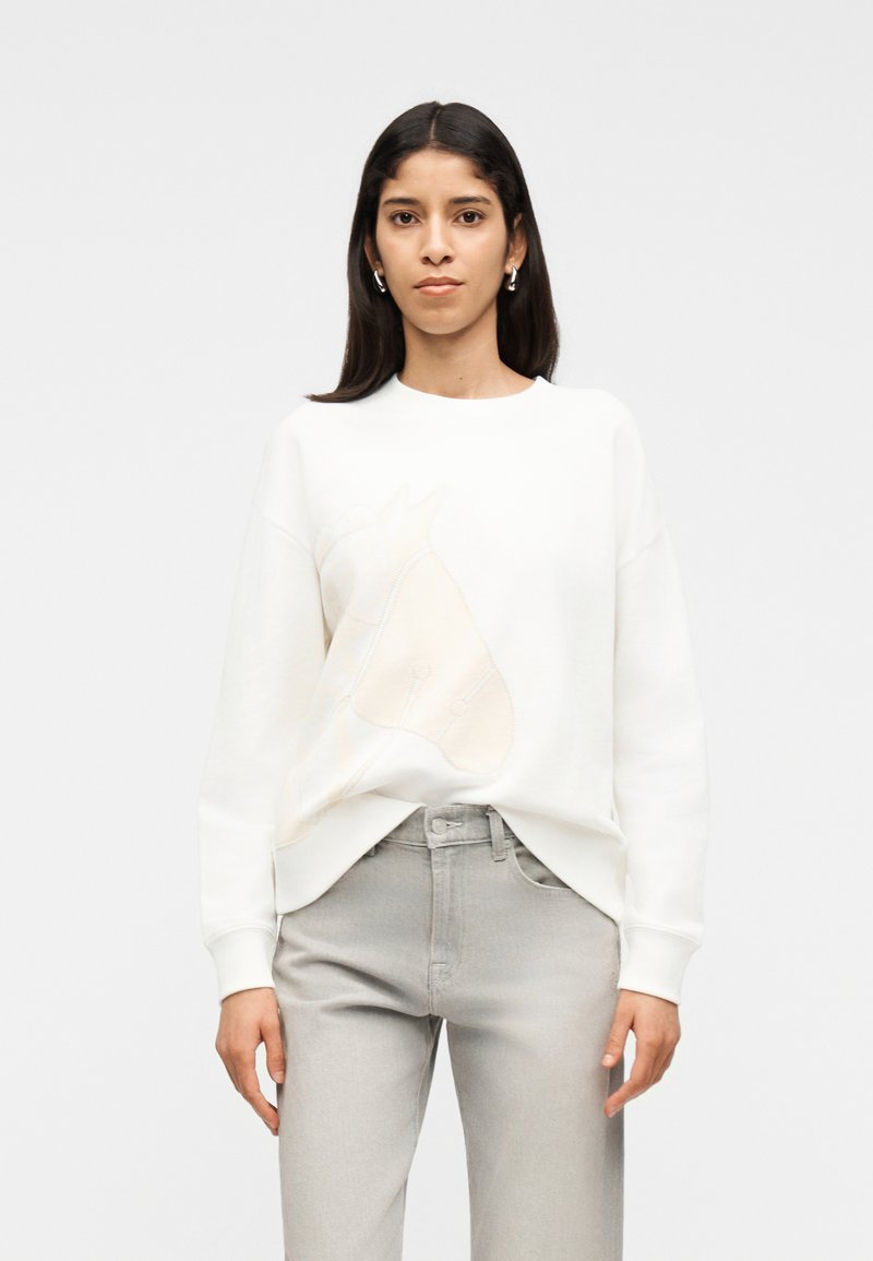 Γυναίκα με μακριά σκούρα μαλλιά που φοράει ασημένια σκουλαρίκια κρίκους, λευκό φούτερ με περίγραμμα αλόγου και ανοιχτό γκρι ψηλόμεσα τζιν, όρθια.