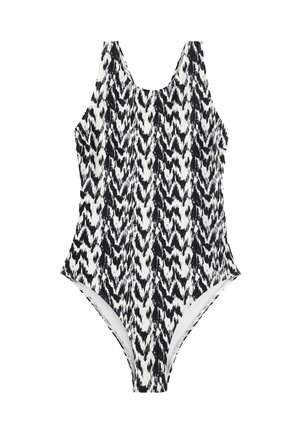 Femme aux cheveux mouillés portant un maillot de bain une pièce à motif noir et blanc, debout sur une plage avec des vagues de l'océan en arrière-plan.