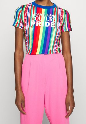 Person trägt ein regenbogen-gestreiftes Versace Jeans Couture Pride T-Shirt kombiniert mit hoch taillierten, leuchtend pinken Hosen vor einem schlichten Hintergrund.