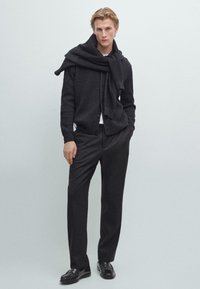 Zwarte geribbelde cardigan over een witte shirt, gecombineerd met donkere broek en zwarte schoenen. De cardigan heeft een brede kraag en een casual gedrapeerde sjaal.