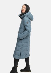 Puhasti, dugi kaput u mekoj plavoj boji, s kapuljačom, džepovima na zatvarač i prošivenim dizajnom. Uparen s crnim hlačama i čvrstim crnim čizmama.