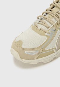 Beige sneaker with mesh and suede materials, reflective accents, textured surface, and a curved toe shape. Laces in a matching tan color.