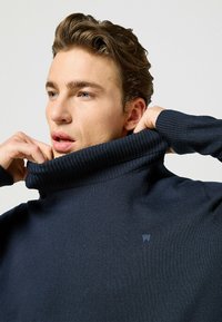 Granatowy sweter z golfem wykonany z prążkowanego materiału. Posiada wysoki kołnierz i subtelny detal z logo na klatce piersiowej, prezentując gładką fakturę.