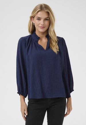 Dunkelblaue Bluse aus strukturiertem Stoff, mit V-Ausschnitt und Rüschensaum. Verfügt über lange Ärmel mit gerafften Manschetten. Kombiniert mit schwarzen Hosen.