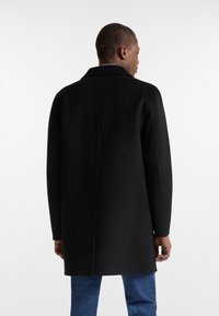 Abrigo negro de lana con corte recto, cuello de muesca y mangas largas. Presenta un diseño limpio en la parte trasera y llega hasta la mitad del muslo.