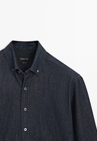 Camicia di jeans con bottoni in blu scuro. Presenta un colletto appuntito, una tasca sul petto singola e bottoni in metallo argento sul davanti e sui polsini.