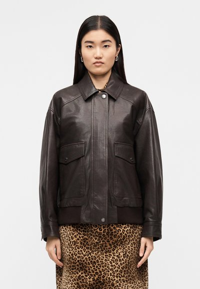Veste en cuir marron avec un col, des poches avant et des poignets côtelés. Portée sur une jupe à imprimé léopard. Texture lisse.
