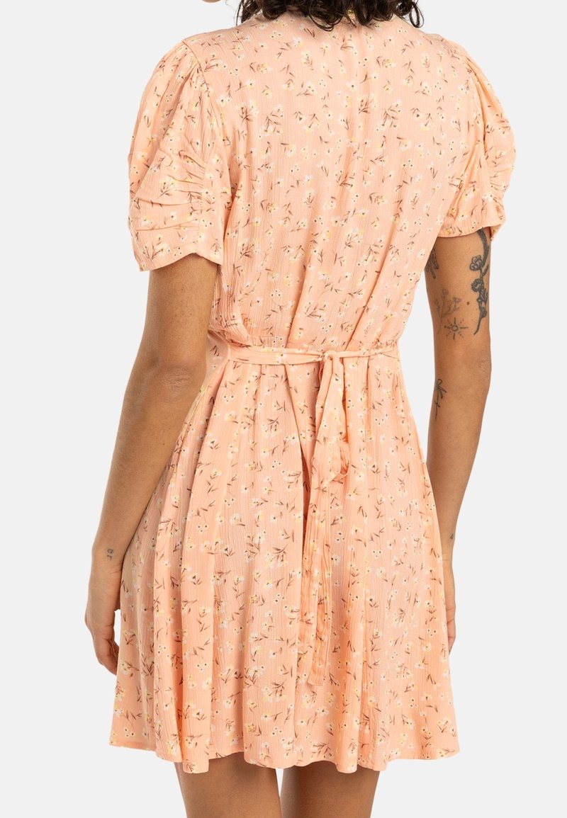Femme portant une robe courte fleurie couleur pêche avec des manches bouffantes et une ceinture nouée, vue de dos, avec des tatouages visibles sur les bras.