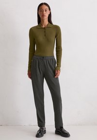 Olijfgroene geribbelde langetermijn polo met kraag, gecombineerd met grijze tapered broek en zwarte instap schoenen, staand tegen een neutrale achtergrond.