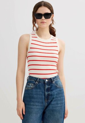 Mujer con gafas de sol negras, camiseta sin mangas blanca con rayas rojas y vaqueros de talle alto azules sobre un fondo liso.