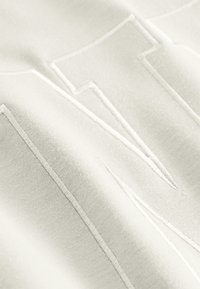 Lichtbeige stof met verhoogde, geborduurde letters in een contrasterende kleur. De textuur is zacht met zichtbare stikdetails.