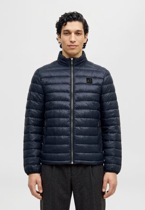 Un homme se tient devant un fond clair et uni, portant une veste matelassée bleu marine foncé avec une fermeture éclair sur le devant et un col montant.