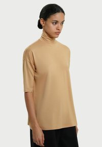 Kortærmet højhalset top i beige stof, med en afslappet pasform og glat tekstur, placeret mod en ensfarvet baggrund.