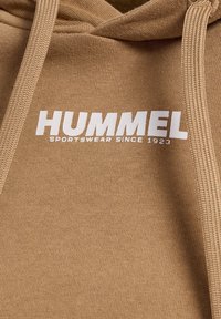 Beige hættetrøje med hvidt "HUMMEL SPORTSWEAR SINCE 1923" logo trykt på brystet, der viser strikket stoftekstur og snore.