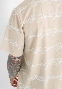 Beige kortermet skjorte med et hvitt håndskrevet mønster av "June Sixth" over stoffet. Myk tekstur med avslappet passform.