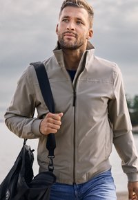 Mann trägt beige Reißverschlussjacke und blaue Jeans, trägt schwarze Umhängetasche, geht draußen in der Nähe von Wasser unter bewölktem Himmel.