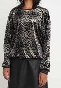 Halenka s leopardím vzorem v černé a šedé barvě z sametového materiálu, volného střihu, s dlouhými rukávy a černým lemováním u výstřihu a manžet.