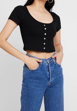 Zwarte geribbelde crop top met korte mouwen, ronde halslijn en zes metalen knopen aan de voorkant; gecombineerd met high-waisted blauwe jeans.