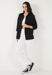 Black button-up shirt with ribbed texture, white crop top, wide white trousers, and black sneakers. Neutral background.