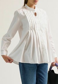 Witte blouse met lange mouwen en een mandarin kraag, met verticale plooien aan de voorkant en een losse pasvorm. Zacht materiaal en knopen aan de voorkant.