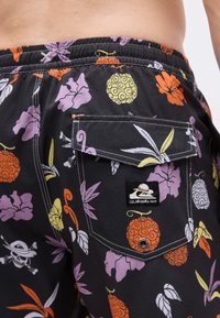 Κοντινό πλάνο μαύρων σορτς με πολύχρωμα λουλουδάτα και στροβιλισμένα φρουτώδη μοτίβα, που διαθέτουν πίσω τσέπη με καπάκι και ετικέτα με το λογότυπο Quiksilver.