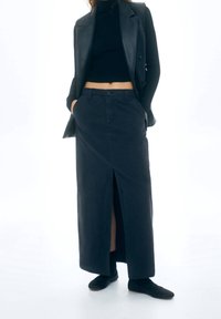 Jupe longue noire avec fente avant, fabriquée dans un tissu lisse. Portée avec un haut noir ajusté et un gilet en cuir, avec des poches latérales et des boutons.