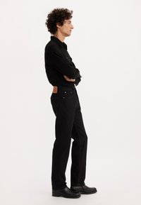 Moški s kodrastimi lasmi stoji v profilu s prekrižanimi rokami, oblečen v črno srajco, črne kavbojke in črne usnjene čevlje na belem ozadju.