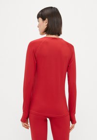 Dlhý rukáv športová blúzka v jednoliatej červenej farbe. Má priliehavý strih bezšvových ramien a zaoblený výstrih. Hladká textúra.