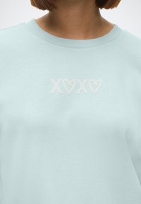 Gaiši zils pusmajci ar ribotu apkakli, centrā ar nodrukātu "XOXO" baltos sirsniņu motīvos. Mīksta tekstūra, brīva piegriezuma.