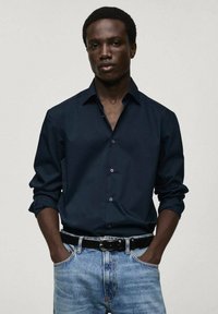 Donker marineblauw overhemd met lange mouwen, zachte textuur, gecombineerd met lichtblauwe denim jeans en een zwarte riem. Eenvoudig, strak ontwerp.