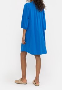 Abito blu, lungo fino al ginocchio, con maniche a palloncino, in tessuto morbido e dal taglio semplice. Indossato con scarpe beige senza lacci, che mostrano uno stile casual.