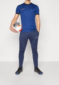 Meriharmaa Nike-urheilupaita lyhyillä hihoilla, yhteensopivat juoksushortsit ja mustat jalkapallokengät. Pelaaja pitää käsissään kuvioitua jalkapalloa.