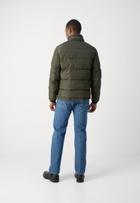 Olive green puffer jacket with a stand collar, horizontal quilting, and long sleeves, paired with light blue denim jeans and black shoes.