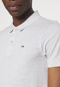 Jasnoszara koszulka polo o teksturowym wykończeniu, z trzema guzikami przy dekolcie oraz małym haftowanym logo w kolorach czerwonym, białym i niebieskim.